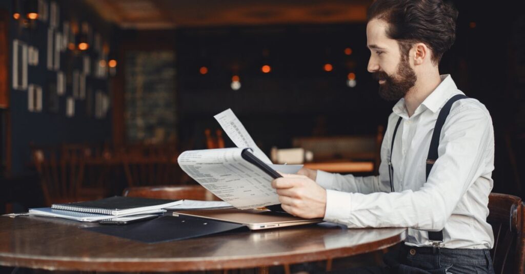 A man with a beard reads a printout while sitting at a wooden table in a cozy café setting.